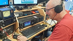 Ein Mann mit Kopfhörern sitzt vor verschiedenen Geräten, die auf einem Schreibtisch stehen.