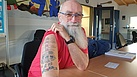 Ein Mann mit Brille und langem weißem Bart hebt den Ärmel seines T-Shirts an. Auf seinem Oberarm ist ein Tattoo, das unter anderem einen Leuchtturm darstellt.